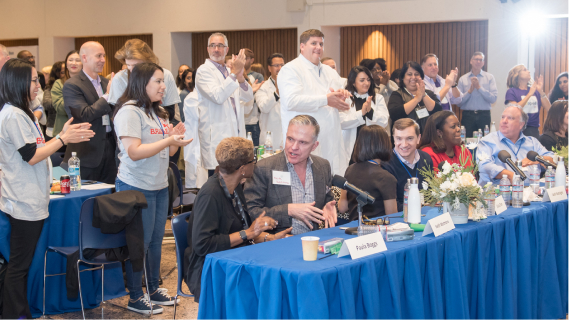 A large group of people fill a convention center room. Some of them are standing and clapping and some are seated and holding discussions with smiles on their faces