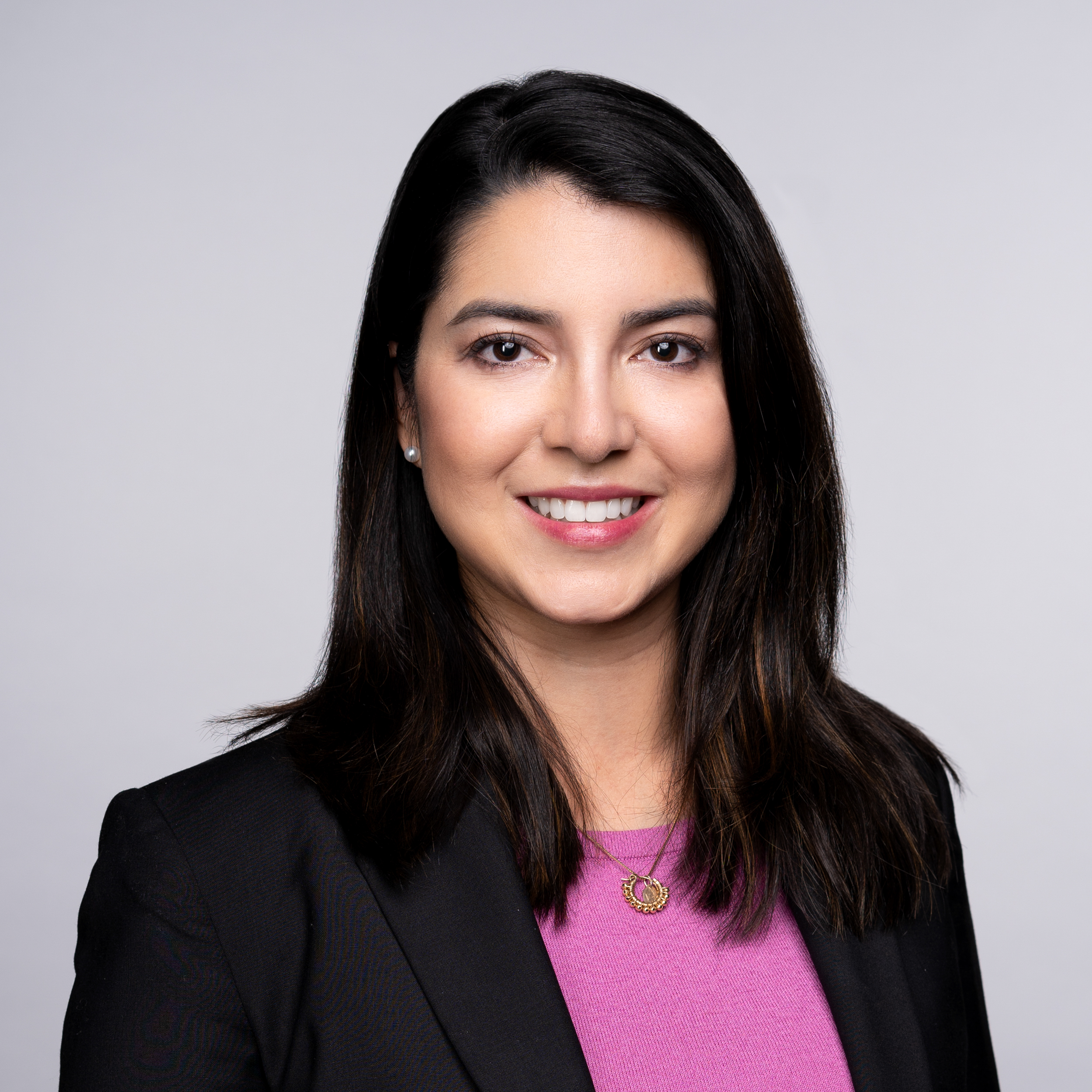 A headshot of Valerie Portilllo, a woman with dark shoulder-length hair and brown eyes wearing a black blazer and a pink top smiling against a gray backdrop