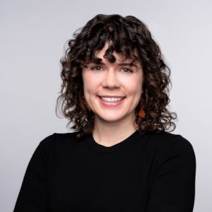 A White woman with curly brown hair smiles in front of a plain gray backdrop.