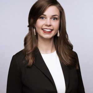 Headshot of Vianna Fagel, a white woman in her 30s with curled brown hair. She is wearing a black blazer and white earrings and is smiling broadly at the camera.