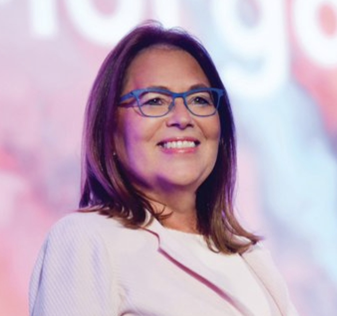 Headshot of Jami McKeon, a woman with glasses and a pink blazer smiling onstage